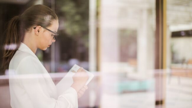 Reflecterende manager maakt aantekeningen bij raam Zakelijke vrouw schrijft geconcentreerd iets op terwijl ze naar buiten kijkt, symbool voor reflectie bij ACT-therapie voor managers.
